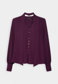 Blouse à manches longues violet foncé avec fermeture à boutons sur le devant et détail d'un nœud en tissu assorti sur fond clair.
