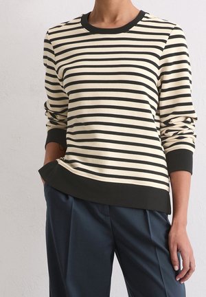 Person trägt ein cremefarbenes und schwarzes Langarmshirt mit horizontalen Streifen, schwarzen Bündchen und Saum, kombiniert mit dunkelblauer Hose.