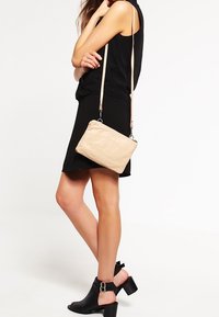 Beige läder crossbody-väska med en texturerad yta, silverfärgad hårdvara och justerbar rem, kombinerad med en svart klänning och ankelskor.