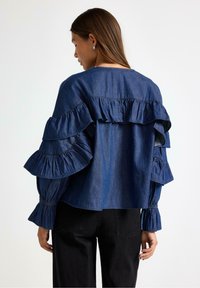 Denimbluse i mørkeblå med lagdelte flæsedetaljer på ærmerne og ryggen. Lange ærmer med elastiske manchetter. Båret med sorte bukser.