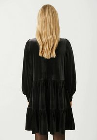 Robe en velours noir avec manches longues bouffantes et design évasé à volants. Texture douce, longueur mi-longue, avec un décolleté arrondi dans le dos.