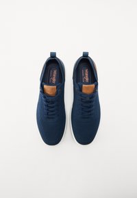 Sapatilhas azul-marinho com parte superior em malha, detalhes em couro castanho na língua e sola de borracha branca. Apresenta um design de atacadores e uma alça de puxar.
