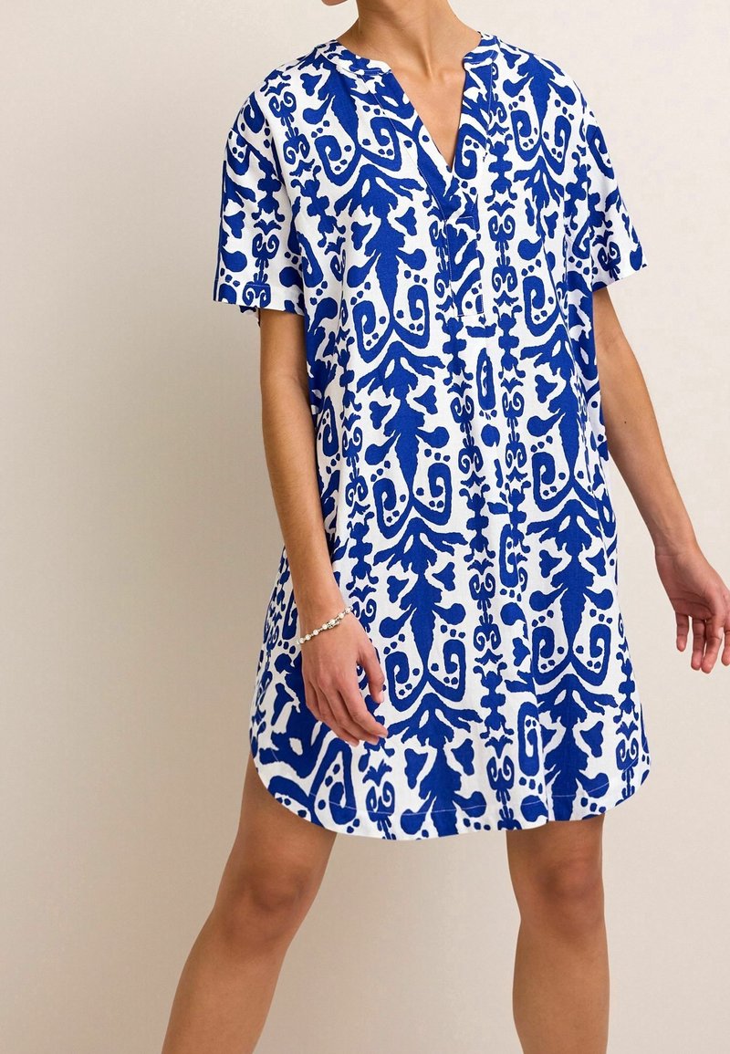 Femme portant une robe blanche à manches courtes, coupe ample, avec des motifs floraux abstraits bleus et un décolleté en V, debout devant un fond uni.