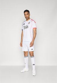 Equipamento de futebol branco com detalhes em vermelho e preto, com o logótipo "Beko". Inclui calções brancos e meias com riscas pretas, combinados com chuteiras brancas.