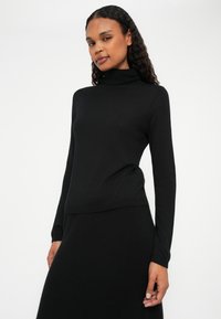 Črna turtleneck puli s dolgimi rokavi, izdelan iz mehkega pletenega materiala. Prilegajoča zasnova z gladko teksturo in diskretnimi rebrastimi detajli na robu.