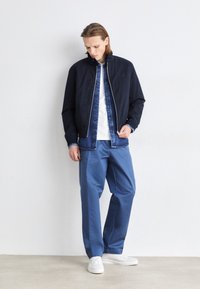 Chaqueta de bombardero azul marino sobre una camisa de mezclilla, combinada con pantalones en un tono azul claro y zapatillas blancas. Texturas suaves y corte relajado.