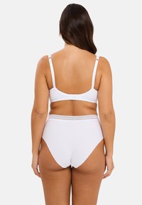 Femme aux longs cheveux noirs portant un soutien-gorge à bretelles ajustables blanches et une culotte taille haute, vue de dos contre un fond uni.