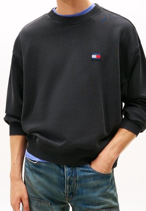 Personne portant un sweat-shirt Tommy Jeans noir avec des poignets et une taille côtelés, une chemise bleue en dessous et un jean bleu en denim, main dans la poche.