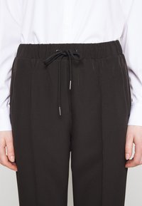 Pantalons noirs avec une taille à cordon, présentant des plis et des poches latérales, fabriqués à partir d'un tissu lisse avec une texture légère.