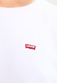 Sudadera blanca con cuello redondo que presenta una pequeña etiqueta roja de Levi's en el área del pecho izquierdo. Textura suave, diseño informal.