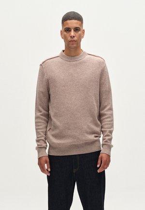 Suéter de punto marrón claro con cuello redondo, puños y cintura acanalados; presenta sutiles aberturas laterales y una textura suave. Usado con jeans oscuros.