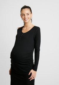 Top de maternidade preto com relevo, mangas longas, decote em forma de barco e detalhes em botão. Apresenta laterais franzidas para maior conforto e ajuste.