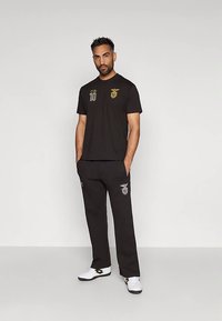 Mees seisab käed taskus, seljas must spordiriietus, millel on kuldne ja valge embleem ning number 10 särgil, valged tossud hallil põrandal.