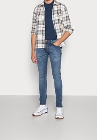 Rutig skjorta i beige, marinblå och brun över en marinblå T-shirt, parat med ljusblå skinny jeans och vita sneakers med svarta detaljer.