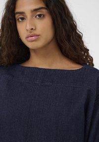 Mladá žena s kučeravými hnedými vlasmi, která nosí tmavomodrý texturovaný top so širokým štvorcovým výstrihom na jednoduchom svetlom pozadí.