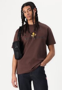 Brun bomulls T-skjorte med et gult tegneseriefigur design og svart tekst. Kombinert med mørke denimbukser og en liten svart crossover-veske.