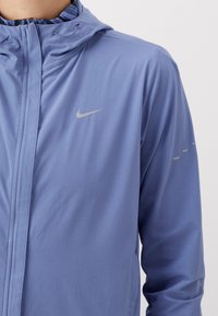 Homme portant une veste à capuche Nike bleu clair avec une fermeture éclair à l'avant et un logo réfléchissant sur la poitrine et la manche, sur un fond uni.