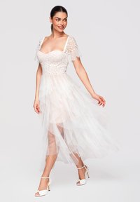Femme portant une robe blanche en dentelle avec une jupe en tulle transparent et des manches courtes en dentelle florale, posant avec la main soulevant la jupe.