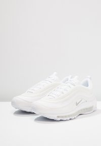 Un paio di sneakers Nike Air Max 97 bianche con ammortizzazione ad aria visibile e loghi swoosh argentati, esposte su una superficie bianca contro uno sfondo grigio.