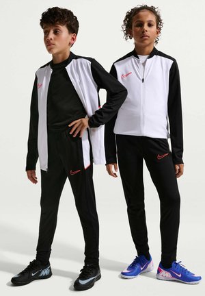 Dwoje dzieci stoi, ubranych w czarno-białe sportowe kurtki i spodnie Nike z czerwonymi logo, czarne i niebieskie buty sportowe Nike na białym tle.