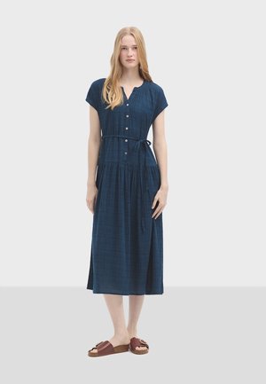 Vestito blu navy con bottoni, maniche corte, vita con cintura e gonna a strati. Indossato con sandali marroni a scivolo con un accentuazione a fiocco.