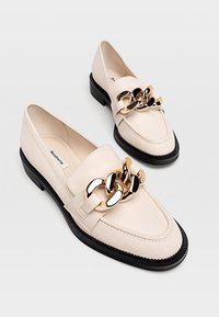 Krémové kožené mokasíny s černou gumovou podrážkou a zlatým a černým řetězovým akcentem přes přední část. Hladký povrch a klasický design.