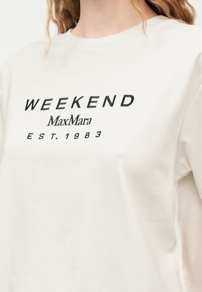 Λευκό πουκάμισο με μαύρο κείμενο που γράφει «WEEKEND MaxMara EST. 1983» φορεμένο από ένα άτομο με κυματιστά ξανθά μαλλιά.