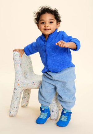 Kleinkind trägt blaue Fleecejacke, hellblaue Fleecehose und blaue Schuhe und steht, während es einen gesprenkelten, mehrfarbigen texturierten Stuhl hält.