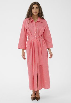 Frau trägt ein rosa langärmliges, tailliertes Kleid mit kleinen gelben Blumenmustern und braune spitze Schuhe, stehend vor einem weißen Hintergrund.