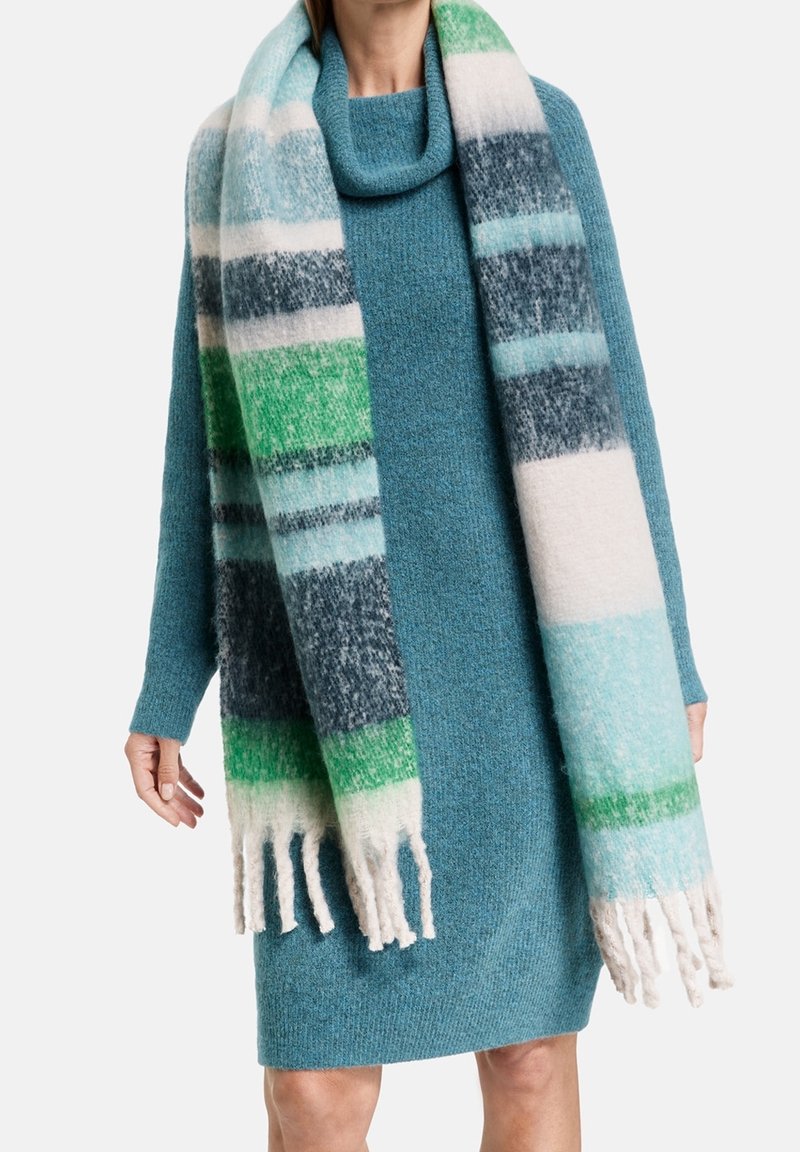 Personne portant une robe-pull turquoise et une longue écharpe duveteuse avec de larges rayures vertes, blanches, bleues et gris foncé avec des franges aux extrémités.