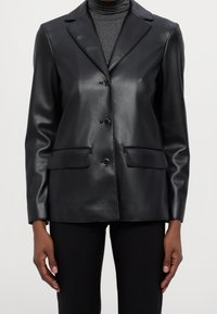 Svart konstläderblazer med kammade slag, enfaldig knappstängning och två framfickor. Slät textur med minimal sömnad.