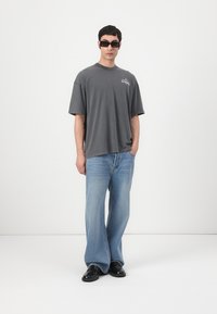T-shirt gris surdimensionné, jeans larges bleu clair, chaussures noires et lunettes de soleil angulaires sombres, présentés sur un fond blanc.