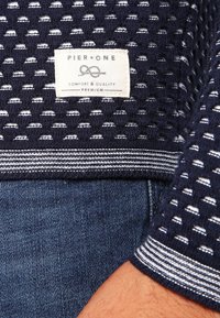 Pull en maille texturée marine avec accents blancs, doté d'une étiquette tissée portant l'inscription "Pier One" et d'une bordure rayée aux poignets.
