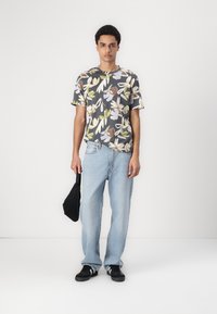 Tricou pentru bărbați cu guler rotund, cu o bază gri închis și un model floral în verde, bej și albastru deschis. Purtați cu blugi decolorati și pantofi negri.