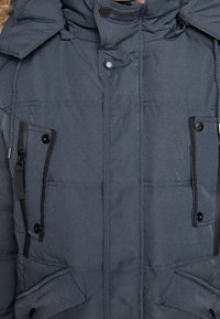 Chaqueta aislante gris oscuro con cuello alto, dos bolsillos en el pecho y cierres frontales de botones a presión. Presenta una tela texturizada y capucha ajustable.