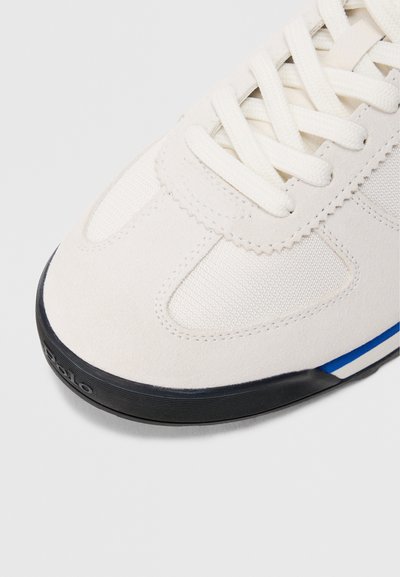 Sapatilha bege com parte superior em camurça e malha, biqueira redonda, atacadores brancos e sola de borracha preta com detalhes em azul e branco.