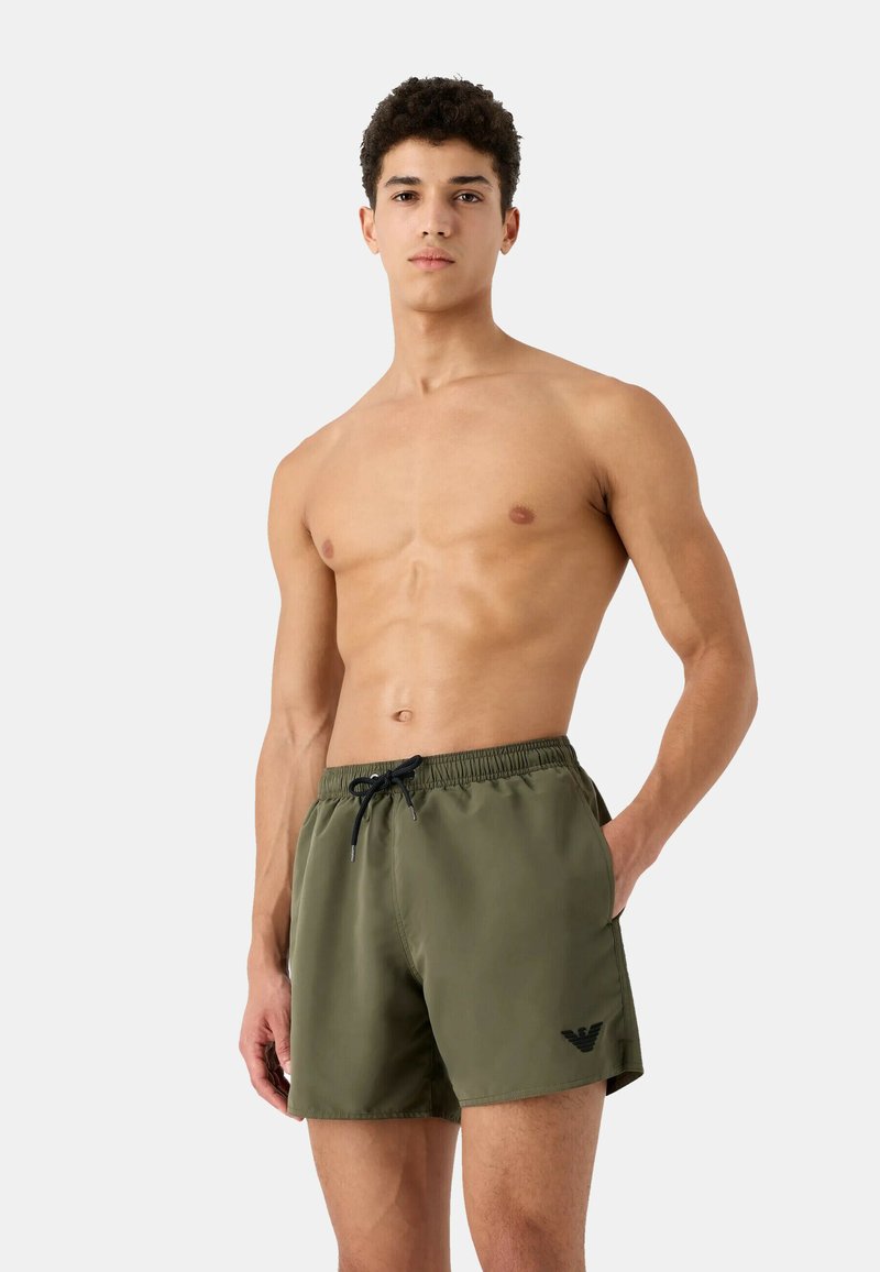 Bañador verde oliva con cintura elástica, cordón ajustable, bolsillos laterales y un logo negro en el dobladillo inferior.