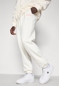 Λευκά παντελόνια φόρμας με κεντημένο λογότυπο "BJÖRN BORG" σε κόκκινο χρώμα, με χαλαρή εφαρμογή, ελαστικούς αστραγάλους και πλαϊνές τσέπες. Συνδυασμένα με λευκά αθλητικά παπούτσια.