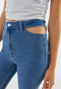 Jeans de mezclilla azul de tiro alto con un detalle de corte lateral, textura suave y diseño clásico de cinco bolsillos. Cierre visible con botón metálico.