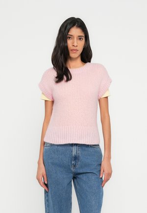 Lyserød strikket sweater med korte ærmer, ribbet kant og løs pasform, lagdelt over en gul skjorte, kombineret med blå denimjeans.