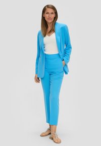 Lichtblauwe op maat gemaakte blazer en bijpassende broek van gladde stof, gecombineerd met een witte top en beige slippers.