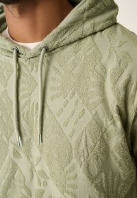 Felpa con cappuccio verde chiaro in tessuto strutturato con motivo in rilievo di forme geometriche e impronte di mani. Dotata di cappuccio con coulisse e texture morbida.