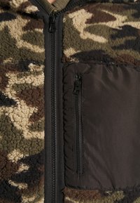 Fleecová bunda na zip v maskáčovém vzoru, s texturovaným béžovo-zeleným povrchem a elegantní černou nylonovou kapsou na zip vpředu.