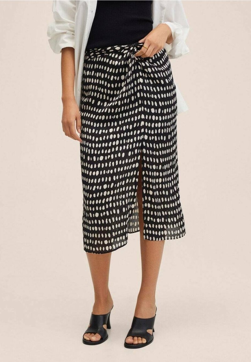Jupe midi à motif noir et blanc avec fente devant, portée avec un top noir, une chemise blanche aux manches retroussées et des sandales ouvertes noires à talons.