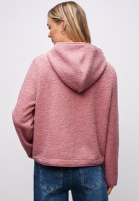 Kobieta z blond włosami ubrana w różowy, teksturowany polar z kapturem i niebieskie dżinsy, widoczna z tyłu na jednolitym tle.