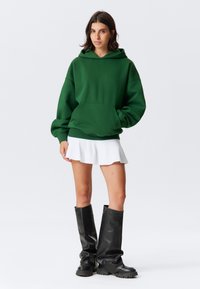Felpa verde oversize con cappuccio e tasca frontale, abbinata a una gonna bianca svasata e stivali neri fino al ginocchio. Sfondo neutro.
