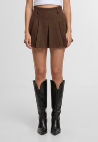 Hnědá plisovaná sukně vyrobená z texturované látky s vysokým pasem a bočními kapsami, v kombinaci s vysokými černými koženými botami s V-čkovým designem.