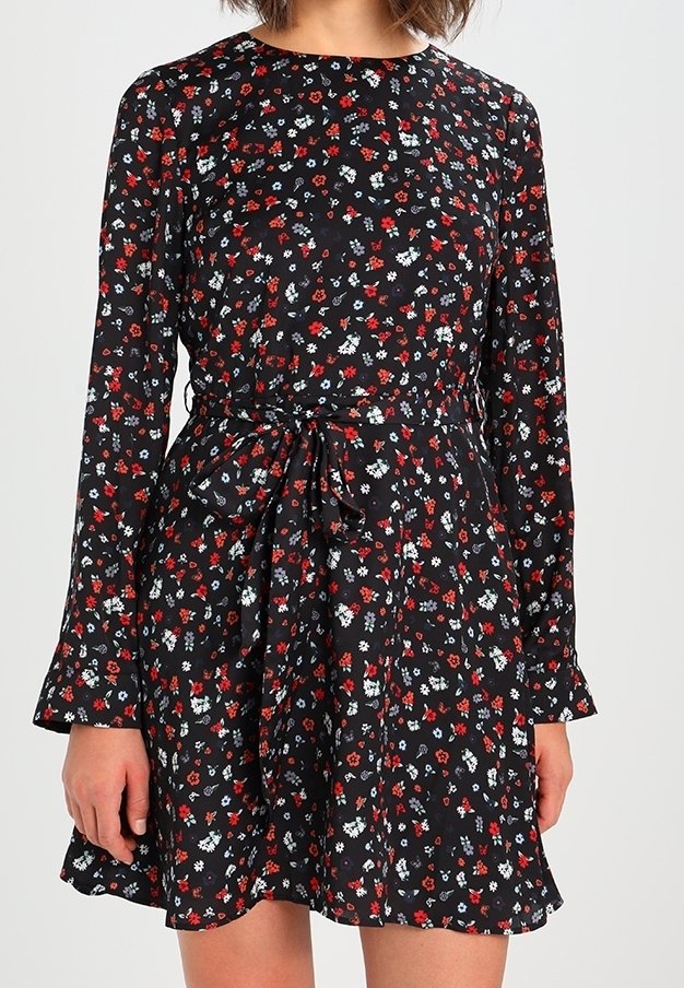 Robe noire à fleurs avec manches longues, ornée de fleurs rouges et blanches, taille cintrée avec un détail noué, et jupe évasée.