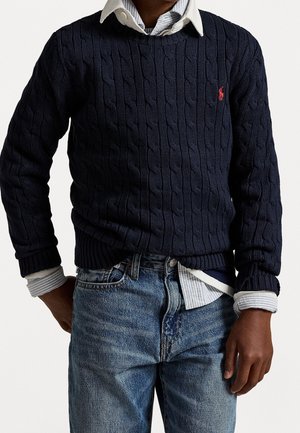 Persona vistiendo un suéter azul marino de punto trenzado sobre una camisa blanca a rayas con cuello y jeans azules, con las manos parcialmente visibles a los lados.