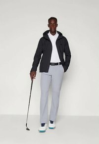 Miestenska športová súprava, ktorá obsahuje čiernu vetrovku, biely polo tričko, šedé nohavice a biele golfové topánky, pričom drží golfovú palicu.